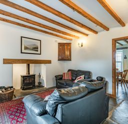 A living room with exposed wooden beams a wood stove two black leather sofas a red patterned rug and a view into a kitchen at Ty Capel Jeriwsalem Bronaber near Trawsfynydd