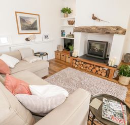 A living room with a beige sofa a fireplace with firewood and a rug at Beudy Hywel in Llanrug near Llanberis