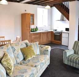 A living room with a floral sofa striped armchair wooden dining table and an open kitchen under wooden stairs at Hendre Aled Cottage 3 in Llansannan