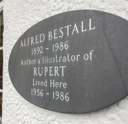 A black plaque on a white wall next to a window commemorating Alfred Bestall author and illustrator of Rupert at Penlan in Beddgelert