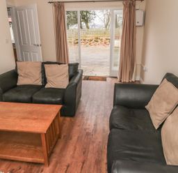 A living room with two black sofas with beige cushions a wooden coffee table wooden floor and glass sliding doors with curtains at No 1 Low Hall Cottages Scalby near Scarborough