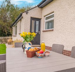 An outdoor table with yellow flowers croissants fruit and orange juice near a house with a black door and windows at Colt Park Cottage in Ulverston