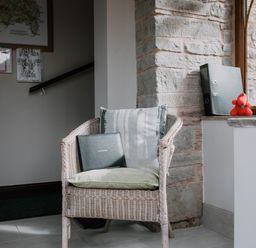 A wicker chair with cushions and a visitors book in a room with a stone wall and window at Stargazer's Field House in Hardwicke near Hay-on-Wye