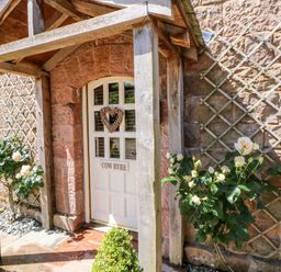 An entrance door with a wooden porch and climbing plants on trellis on the stone wall at The Cow Byre in Barras near Kirkby Stephen