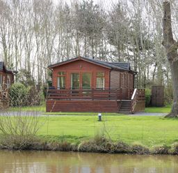 A wooden lodge with a deck and outdoor furniture near a pond with trees in the background at Oak Lodge in Beaconsfield Holiday Park
