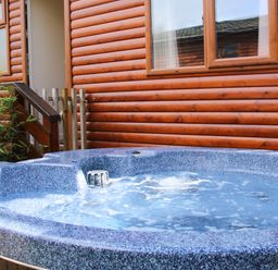 An outdoor hot tub with bubbling water beside a wooden cabin with windows and a small fence