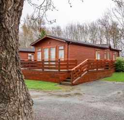 A wooden cabin with a deck and stairs surrounded by grass and trees at Oak Lodge in Beaconsfield Holiday Park