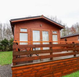 A wooden lodge with a deck railing and outdoor chairs at Oak Lodge in Beaconsfield Holiday Park