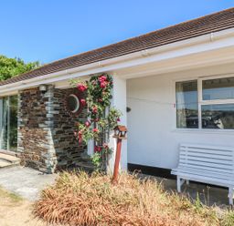 The exterior of a house with a stone and white wall a window a sliding glass door a white bench and a rose bush at The Shack in Mawgan Porth near Newquay