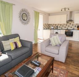 An open living room and kitchen space with a gray sofa and armchair with cushions a glass coffee table and white kitchen cabinets at Middle Barn in Launceston