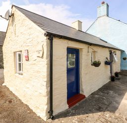 An exterior view of a small cream stone cottage with a blue door and hanging flower pots at Ffynnon Tom in Trefin near St Davids