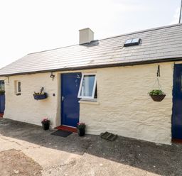The exterior of a single-story building with white stone walls blue doors hanging flower baskets and small potted plants at Ffynnon Tom in Trefin near St Davids