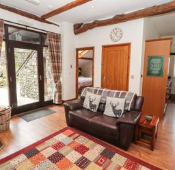 A living room with a leather sofa and plaid pillows next to wooden doors and large glass doors with plaid curtains at The Hayloft in Newlands near Braithwaite and Keswick