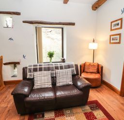A sitting area with a leather sofa with two plaid cushions a brown round chair a floor lamp and a window with a vase of flowers at The Hayloft in Newlands near Braithwaite and Keswick