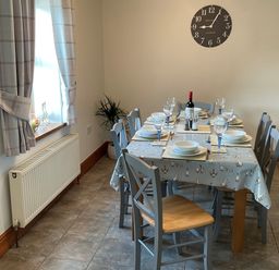 A dining room with a table set for six near a window with curtains and a wall clock at Tangaer Cottage in Cellan near Lampeter