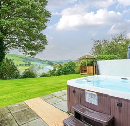 An outdoor hot tub on a stone patio next to a grassy lawn with a wooden gazebo and hills in the background at Old Rectory Cottages Aberhafesp near Newtown