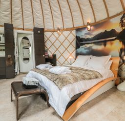 A bedroom with a double bed covered in white linens and a fur throw a small kitchenette area and a glass side table with a lamp at Island Yurt Beckford near Tewkesbury