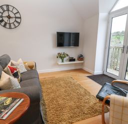 A living room with a gray sofa with cushions a wall clock a wall-mounted TV and glass doors leading outside at The Loft at Lucott House in Bath