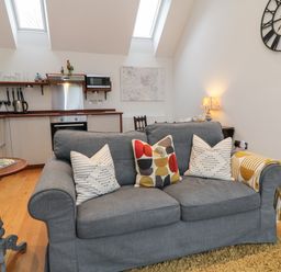 A living room with a grey sofa four pillows a side table and an open kitchen at The Loft at Lucott House in Bath