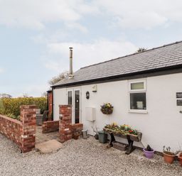 An exterior of a white cottage with a gray tiled roof and small brick patio with outdoor chairs surrounded by flower pots at The Old Stable in Mold