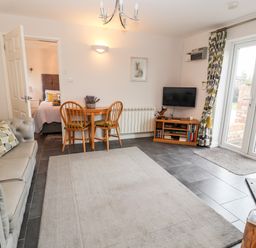 A living room with a gray sofa and patterned cushions a wooden table with two chairs a small wooden TV stand with a television and glass doors leading outside at The Old Stable in Mold