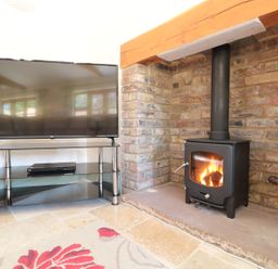 A television on a glass and metal stand next to a wood burning stove with a fire in a room with brick walls and tiled floor at The Old Dairy in Newnham-on-Severn