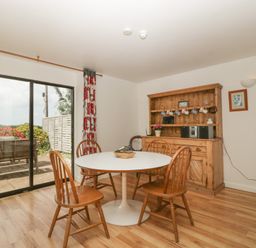 A dining area with a round white table surrounded by wooden chairs and a wooden cabinet with hanging mugs at Wheelbarrow Cottage in Stoke Prior near Leominster