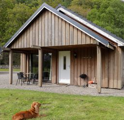 A wooden cottage with a patio table and chairs a barbecue and a dog lying on grass at Stone Water Cottage in Strathpeffer