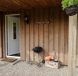 A wooden porch with a white door a small charcoal grill a wicker basket with firewood tools and a hanging basket of flowers at Stone Water Cottage Strathpeffer