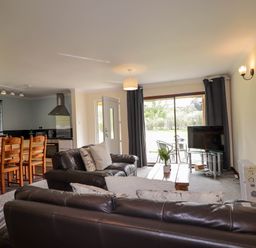 A living room with leather sofas a wooden dining table and chairs a kitchen area and a television near a sliding glass door at Stone Water Cottage in Strathpeffer