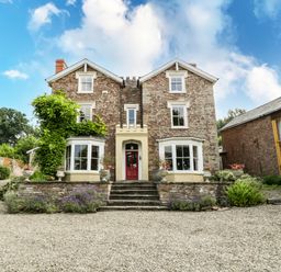 A two-story stone house with a red door and two bay windows surrounded by plants and gravel driveway at Wheelbarrow Castle in Leominster