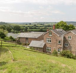 A stone house with multiple chimneys and white windows surrounded by green fields and trees at Wheelbarrow Castle in Leominster