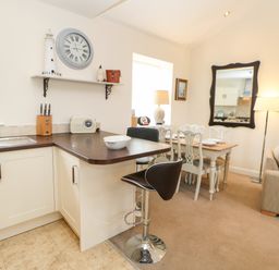 A kitchen with a breakfast bar and stools next to a dining table with chairs and a mirror on the wall at Driftwood Apartment in Amble-by-the-Sea