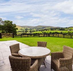 An outdoor patio with wicker chairs and a round glass table overlooking green fields and hills at Beudy in Llangwm near Bala