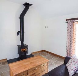 A living room with a black wood stove chimney a wooden table a dark sofa with floral cushions and a door with floral curtains at Beudy in Llangwm near Bala