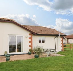 A single story house with a tiled roof green potted plants a picnic table and grass at Broom Cottage in Stoneleigh Holiday Village near Sidmouth