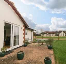 An outdoor patio area with green potted plants sliding glass door and picnic table outside a house at Broom Cottage Stoneleigh Holiday Village near Sidmouth