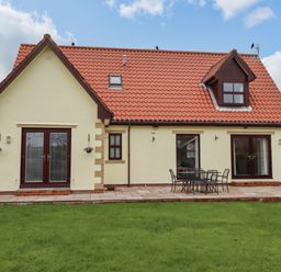 The back view of a house with a red tiled roof a patio with a table and chairs and a green lawn at Darling Cottage in Bamburgh