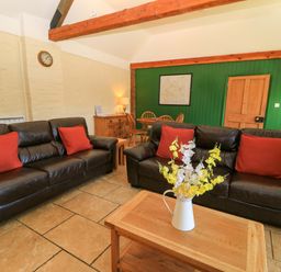 A living room with two leather sofas with red cushions a wooden coffee table with flowers and a dining table in the background at Keepers Cottage in Winterborne Stickland