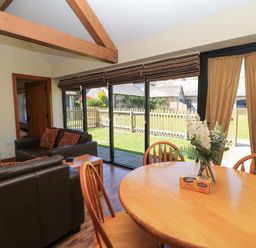 A living room with leather sofas, a wooden dining table with chairs, a vase with flowers, and large glass doors overlooking a garden at New Barn in Winterborne Stickland