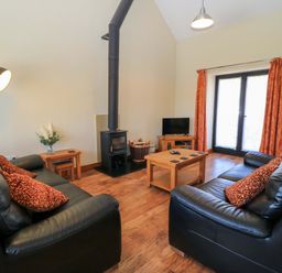 A living room with two black leather sofas, a wood-burning stove, a wooden coffee table, a floor lamp, a small TV on a wooden stand, and glass doors with orange curtains at The Old Cart Shed in Winterborne Stickland
