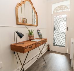 A hallway with a wooden console table holding a lamp plant and brochures below a gold-framed mirror and a glass door with a diamond pattern at The Robins in Filey