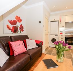 A living area with a brown leather sofa with cushions and a wooden table with a vase of purple flowers and a closed book in a kitchen space at Church View in Ambleside