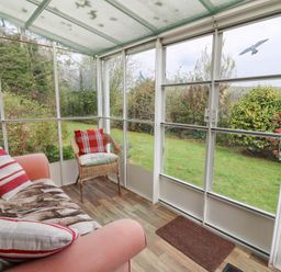 A sunroom with a sofa, cushion, wicker chair, and a view of a garden at Bryn yr Haf in Llanfair Clydogau near Lampeter