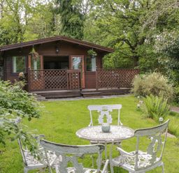 A wooden cabin with a porch and lattice railing in a garden with a white metal table and chairs surrounded by trees at Y Caban in Llechwedd The Conwy Valley