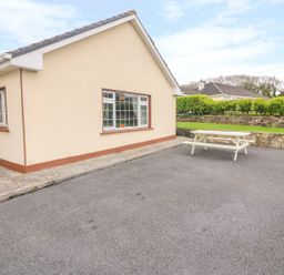 An exterior view of a cream colored building with a window and a door next to a paved area with a white picnic table and green hedges at Island View in Ardmore County Waterford