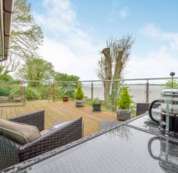 A wooden deck with wicker chairs and a glass table with a coffee press overlooking a glass railing and a coastal view at Solway Cottage in Bowness-on-Solway near Wigton