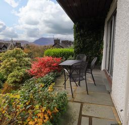 A patio with a table and four metal chairs surrounded by various plants and shrubs with rooftops and mountains in the background at Walkers Retreat Ambleside