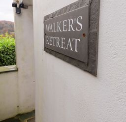 A sign reading Walkers Retreat mounted on a white exterior wall with greenery and hills in the background at Walkers Retreat in Ambleside