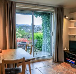 A dining area with wooden table and chairs next to sliding glass doors overlooking a patio with outdoor chairs and greenery in Walker's Retreat Ambleside